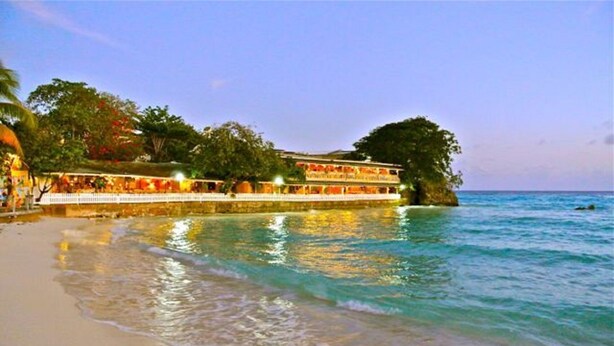 A beachfront hotel with a long structure extending into the water, surrounded by trees and a clear sky.