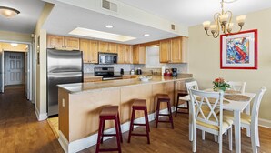Una cocina con armarios de madera, un refrigerador de acero inoxidable, un microondas y una zona de comedor con sillas y taburetes blancos.