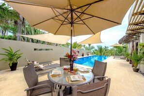 A round table with chairs and a large umbrella, set against a poolside backdrop with potted plants and a clear blue sky.