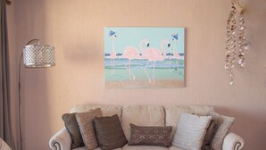 A living room with a beige sofa, patterned cushions, a wall-mounted painting of flamingos, and a hanging shell decoration.