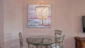 A dining area with a round glass table and metal chairs, a chandelier, and a painting of a beach scene on the wall.