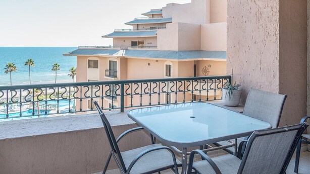 A balcony with a table and chairs, overlooking a pool and a building with multiple balconies.
