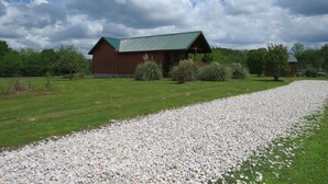 A wooden cabin with a green roof, surrounded by a gravel path and green grass, with trees and shrubs in the background. - SUNDANCER CABIN CADDO RIVER!!!!!   GREAT GETAWAY !!!!!!!! River Access!! (Glenwood)