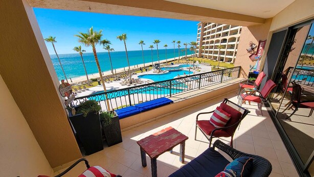 Varanda com vista para uma piscina e praia, equipada com cadeiras e uma mesa.