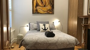 Ett sovrum med en säng, ett nattduksbord, en lampa, en garderob i trä och en tavla på väggen.