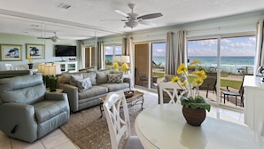 A spacious living room with a large window overlooking the ocean, a ceiling fan, and a dining area with a table and chairs.