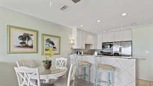 A kitchen with white cabinets, a stainless steel refrigerator, and a dining area with white chairs and a glass table.