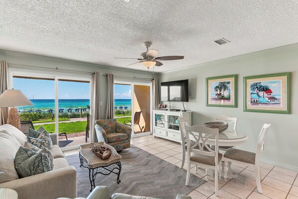 A spacious living room with a view of the ocean, a ceiling fan, and a dining area with white chairs and a glass table.