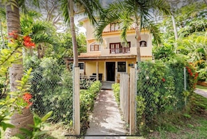 A two-story house with a balcony, surrounded by lush greenery and a pathway leading to the entrance.