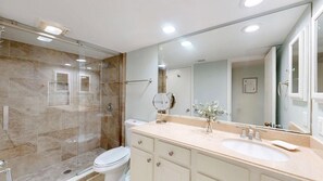 Un baño moderno con cabina de ducha de vidrio, inodoro blanco, tocador color beige con lavamanos y un espejo grande.