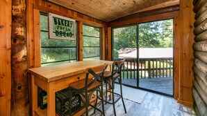 A wooden cabin interior with a sign reading "BAKERY FRESH BREAD," a wooden table, two chairs, and a view of the outdoors through large windows. - Dupont Forest Trailside Cabin (Hendersonville)