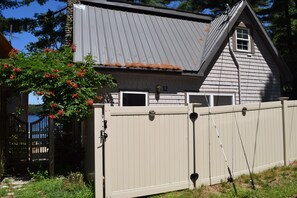 Two-story house with a gabled roof and dormer window, set amidst lush green surroundings and a fence.
