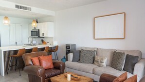 Een moderne woonkamer met een bank, salontafel en stoelen, een keuken met witte kasten en een groot schilderij in een lijst aan de muur.
