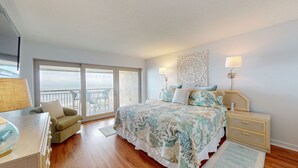 A bedroom with a large bed, a nightstand, a dresser, a chair, and a view of the sea through a sliding door.