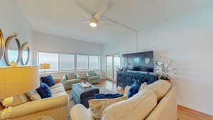 A spacious living room with a large flat-screen TV, comfortable seating, and a view of the ocean through large windows.