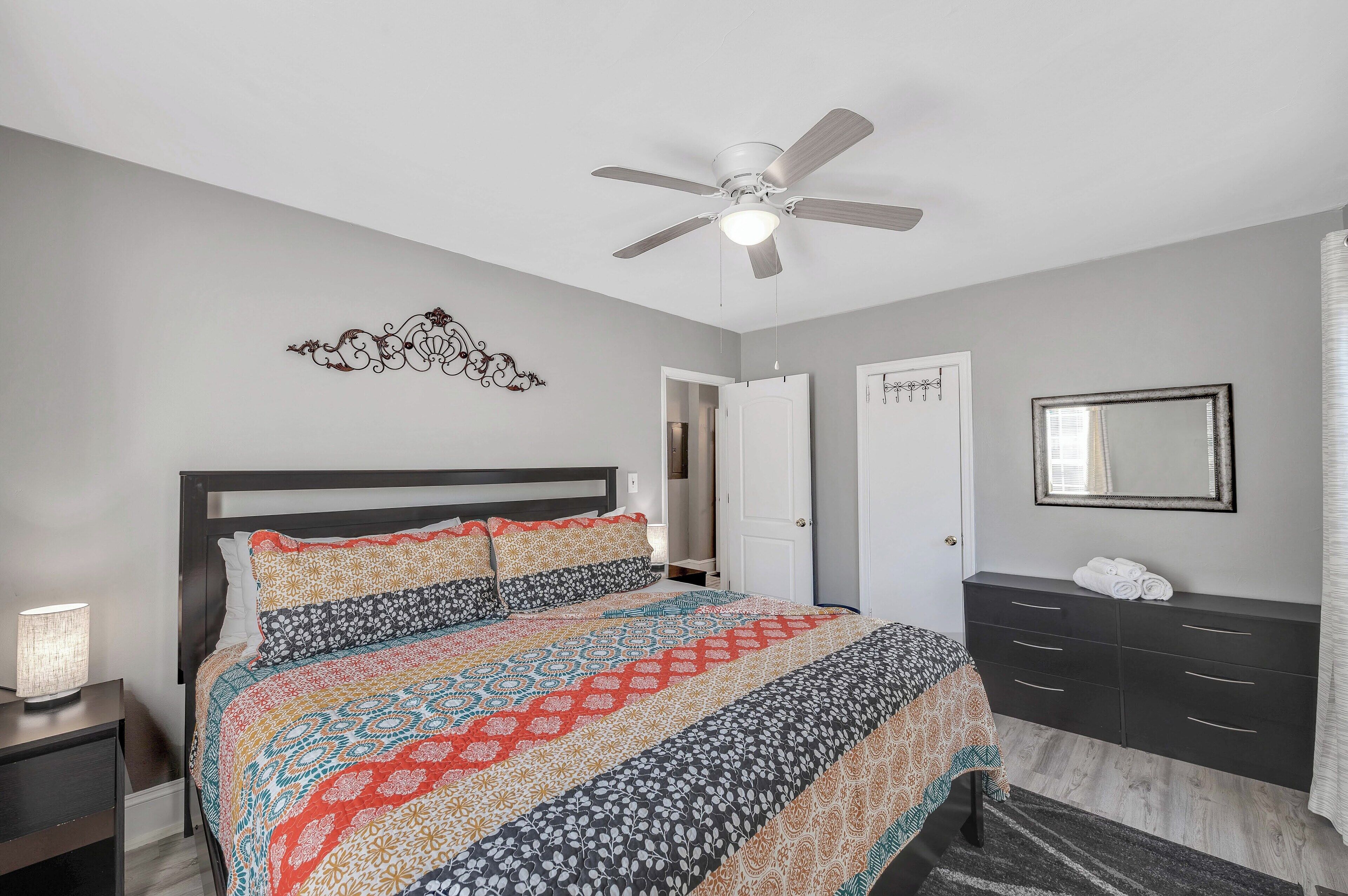 A bedroom with a bed, a ceiling fan, a bedside table with a lamp, a dresser, and a door.