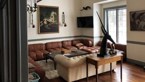 A living room with a brown leather sectional sofa, a beige ottoman, a wooden coffee table, and a mounted deer head. There is a painting of a horse on the wall, a television, and a window with a view of trees. - Greystone Manor - An isolated 30 Acres in Stowe,  with Hot Tub!!! (Stowe)