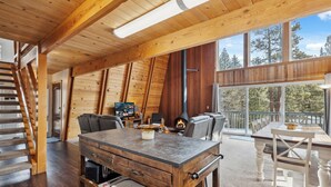 Un acogedor interior de cabaña con una mesa de comedor de madera, asientos cómodos, una chimenea y grandes ventanales que dan a vistas del bosque circundante.