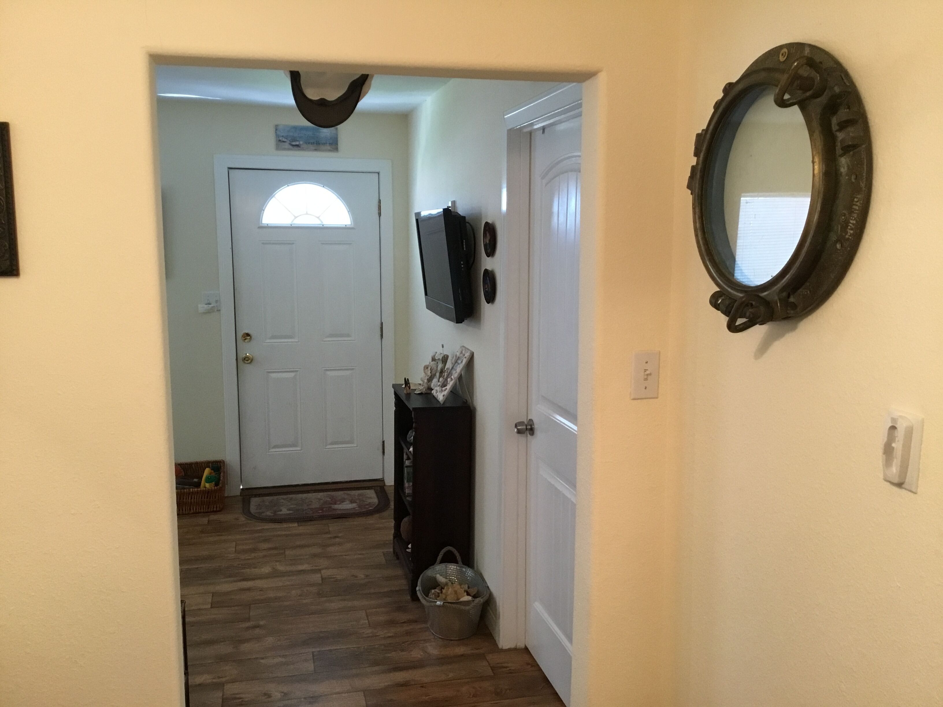 A hallway with wooden flooring, a white door with a semi-circular window, a dark-colored console table with decorative items, a metal bucket, and a wall-mounted mirror.