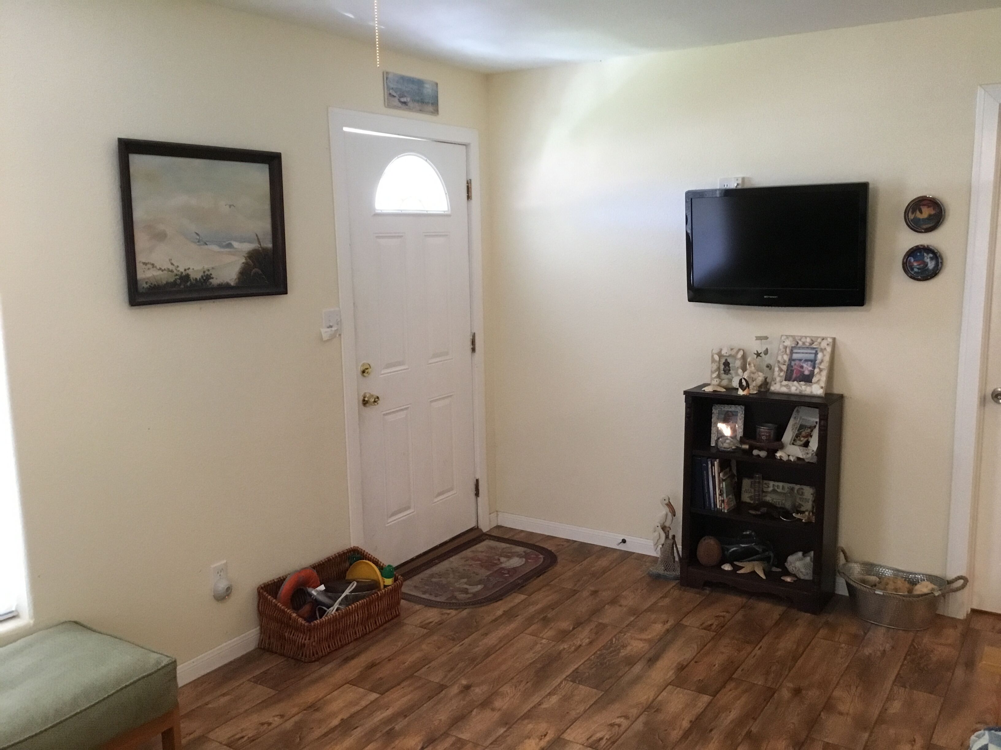 Una habitación con piso de madera, una puerta blanca, un televisor montado en la pared, un pequeño estante con fotos enmarcadas y una cesta de objetos cerca de la puerta.