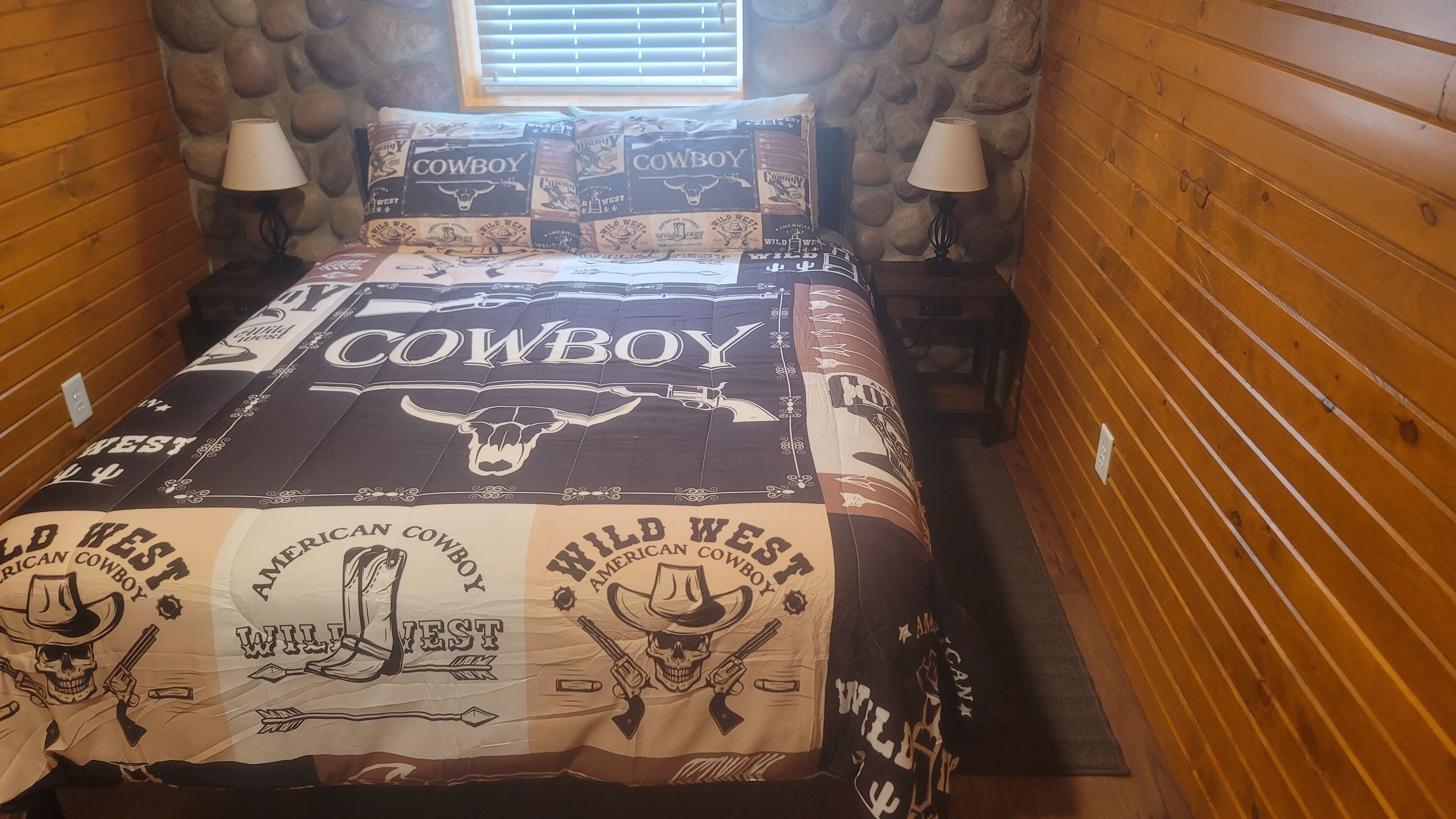 A bed with a 'Cowboy' themed bedspread, two lamps, and a stone wall.