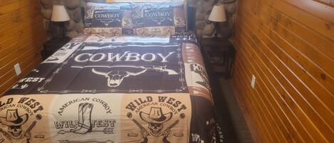 A bed with a 'Cowboy' themed bedspread, two lamps, and a stone wall.