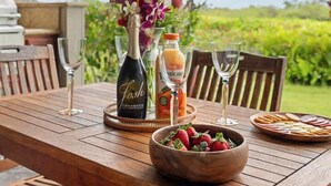 A wooden table set with a bottle of wine, glasses, a bowl of strawberries, and a fruit drink. - Gated Condo with Exclusive Mauna Lani Beach Access (Kamuela)