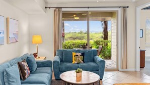 A living room with two blue sofas, a coffee table, and a lamp. There is a sliding glass door leading to a balcony with a view of greenery. - Gated Condo with Exclusive Mauna Lani Beach Access (Kamuela)