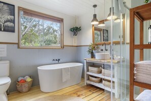 A bathroom with a freestanding bathtub, a large window with a view of trees, a sink with a mirror, and wicker baskets on shelves. - Mandala Bruny Island, waterfront, near Restaurant (Alonnah)