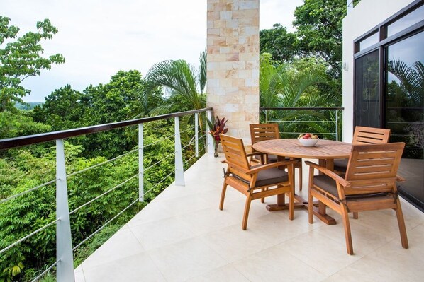 A balcony with a round wooden table and chairs, surrounded by lush greenery.