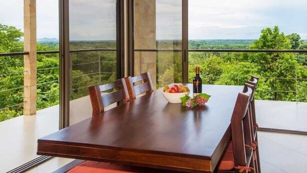 Una mesa de comedor con sillas, un cuenco de fruta y una botella de vino en un balcón con vistas a la vegetación y montañas.