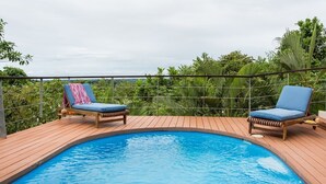 Una piscina con dos sillas reclinables, rodeada por una terraza de madera y una barandilla de metal.