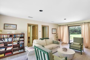 A living room with a sofa, a coffee table, a bookshelf, and a view of a garden through a window.