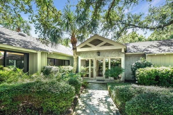 A residential property with a covered patio, a pathway leading to the entrance, and lush greenery surrounding the house.
