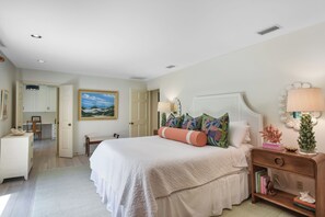A bedroom with a large bed, a bedside table with a lamp, a bookshelf, and a painting on the wall.