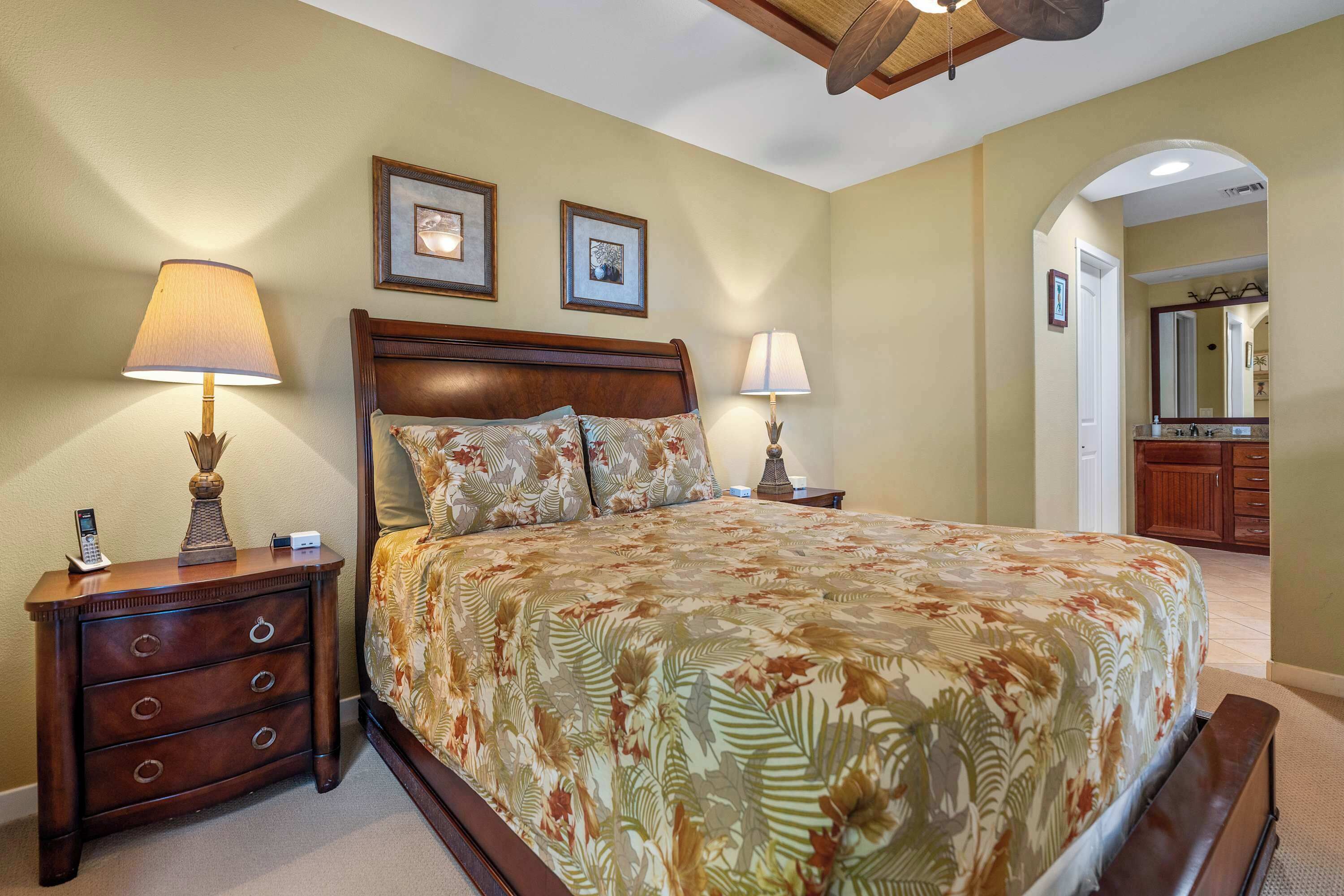 A bedroom with a large bed, two bedside tables, and a nightstand. There are framed pictures on the wall, a mirror, and a door leading to another room.