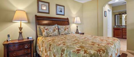 A bedroom with a large bed, two bedside tables, and a nightstand. There are framed pictures on the wall, a mirror, and a door leading to another room.