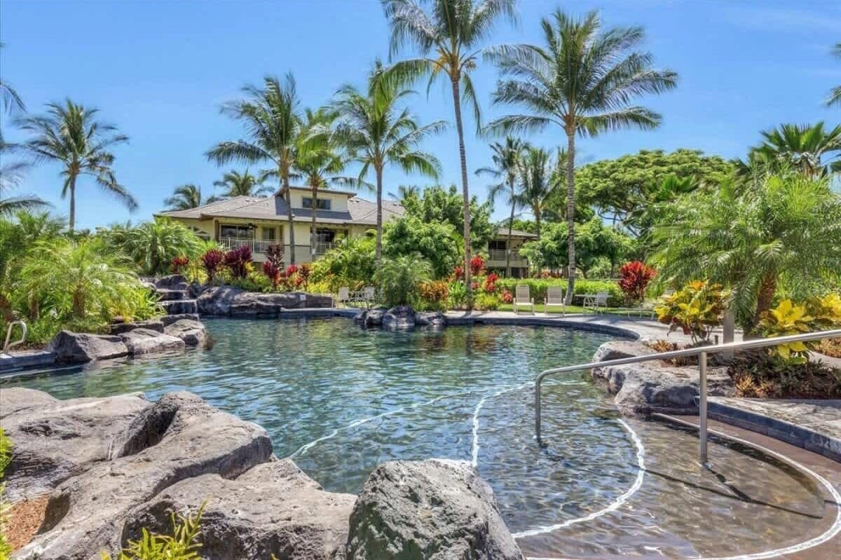 A pool surrounded by rocks and palm trees, with a building in the background.