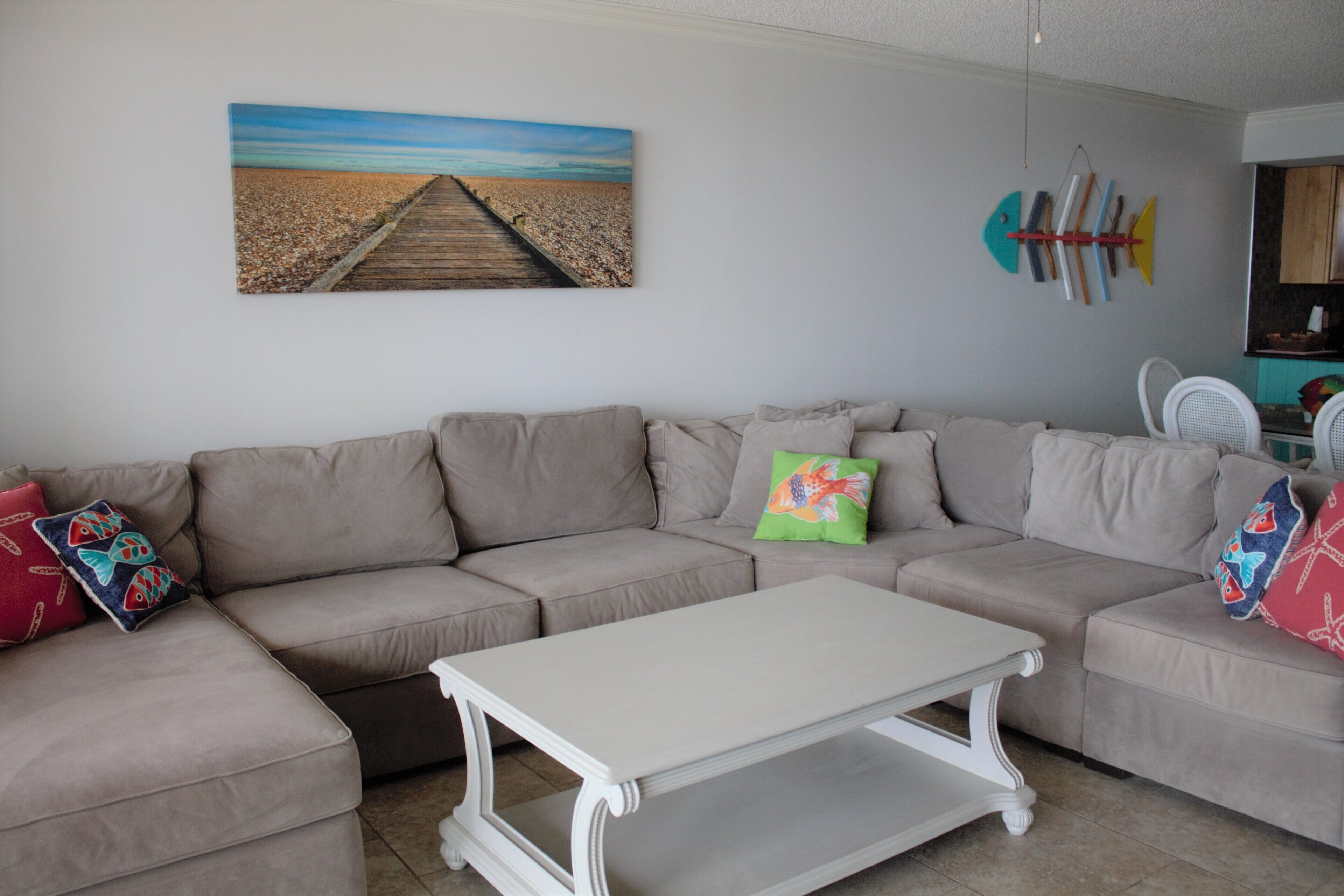 Un salon avec un canapé d’angle beige, une table basse blanche et un tableau représentant une scène de plage.