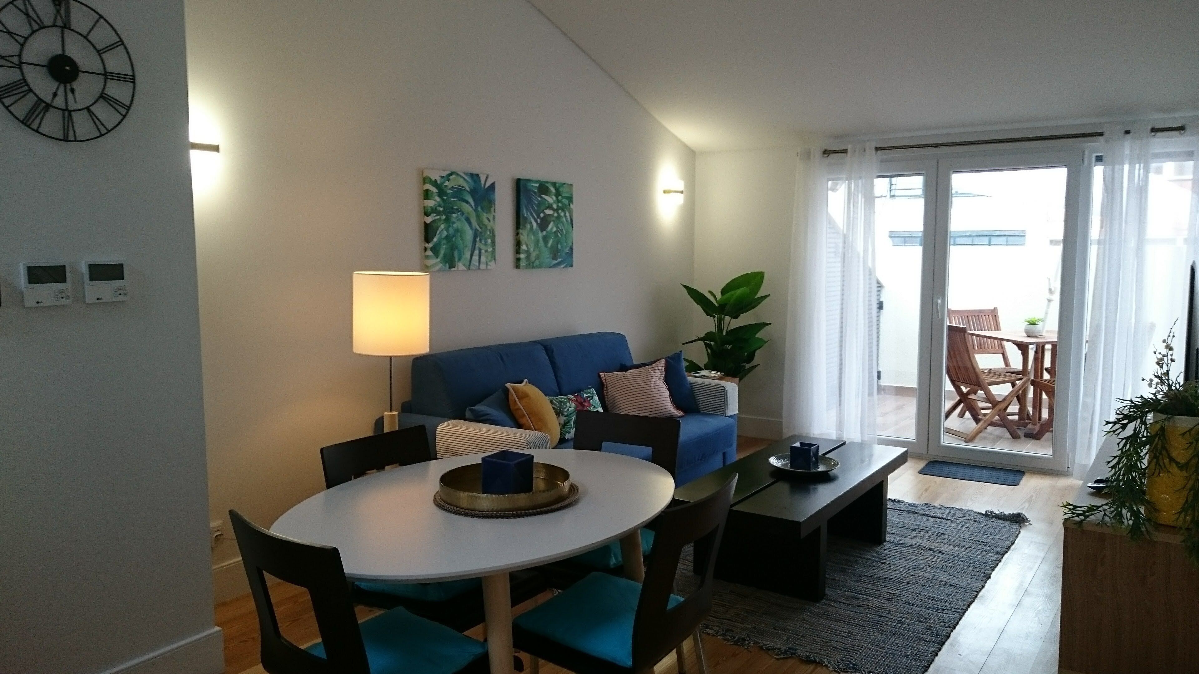 Ein Wohnzimmer mit rundem Tisch, blauem Sofa und einem großen Fenster mit Vorhängen.