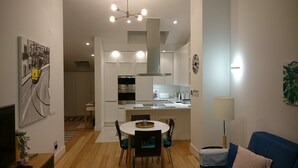 Eine moderne Küche mit Edelstahlgeräten, ein Essbereich mit rundem Tisch und Stühlen sowie ein Wohnzimmer mit Sofa und Lampe.