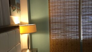 A hotel room with a bed, a nightstand with a lamp, a wall-mounted air conditioner, and a window with blinds.