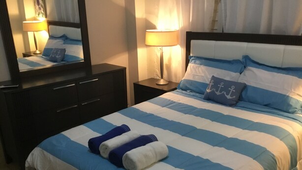 A hotel room with a bed featuring blue and white striped bedding, a nightstand with a lamp, a dresser with drawers, and a mirror on the wall.