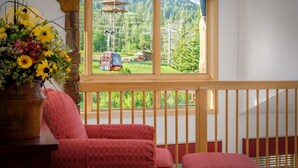 A room with a wooden balcony, a window with a view of a cable car and forest, and a red patterned sofa. - RMR: Cody House C Condo in Teton Village (Teton Village)