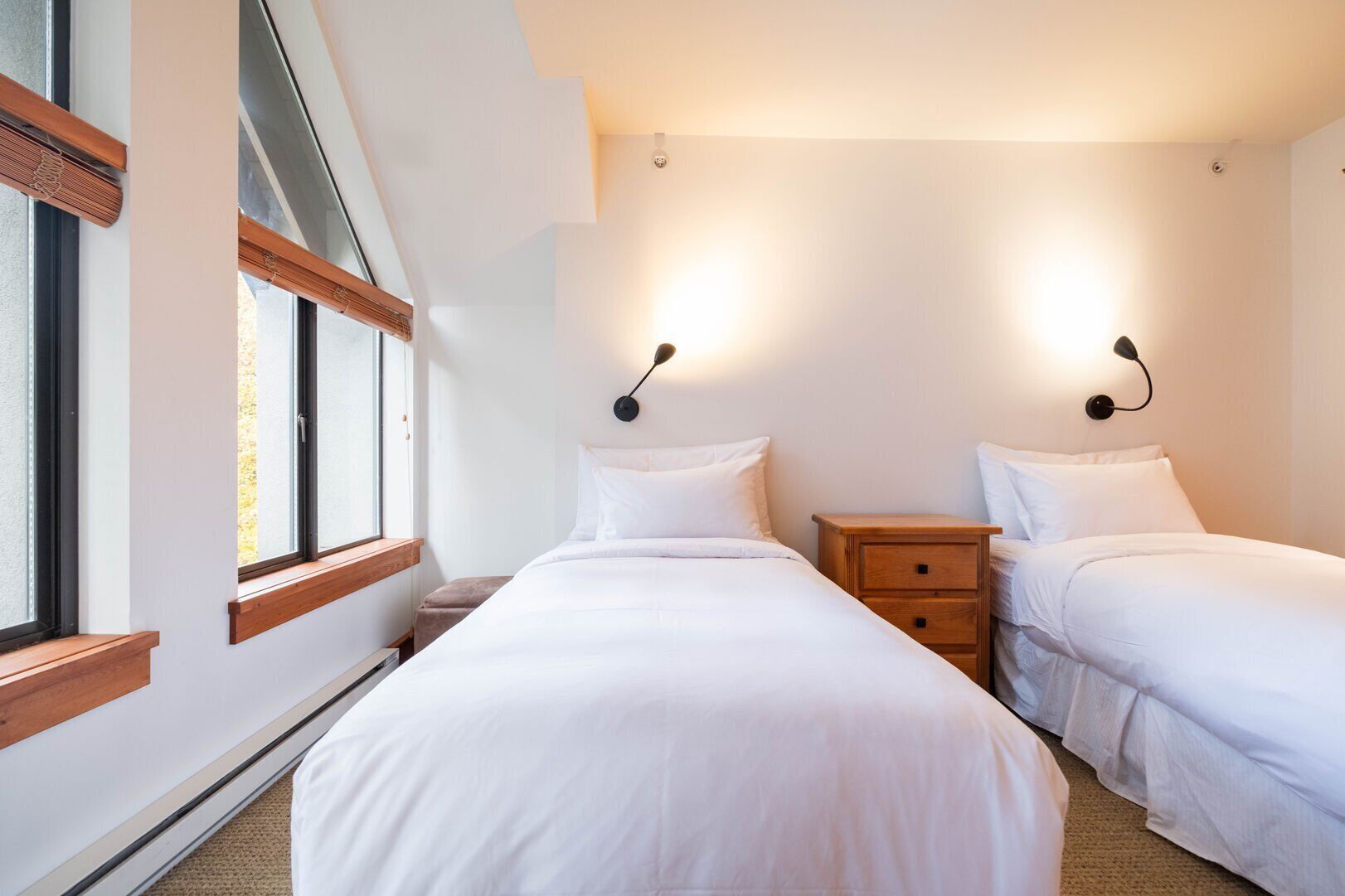 A bedroom with two beds, a wooden nightstand, a window, and wall-mounted lights.