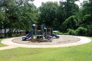 Children can play on the playground's slides and climbing elements in a secure, paved area surrounded by trees.