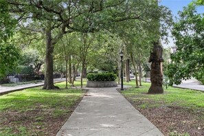 Stroll along the serene park pathway lined with mature trees and a central planter, featuring a historic statue.