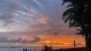 Uma praia com pôr do sol, palmeiras e algumas pessoas caminhando.