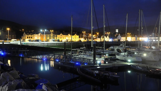 Una marina de nit amb vaixells amarrats, un pont i edificis al fons.
