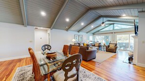 A spacious living area with a dining table, chairs, and a sofa. The room has wooden floors, a high ceiling with exposed beams, and large windows providing natural light. - Golf course and lagoon views! (Hilton Head)